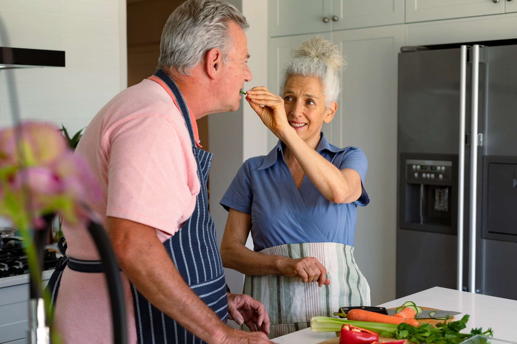 Happy caucasian senior couple standing in kitchen, preparing meal together and eating vegetables