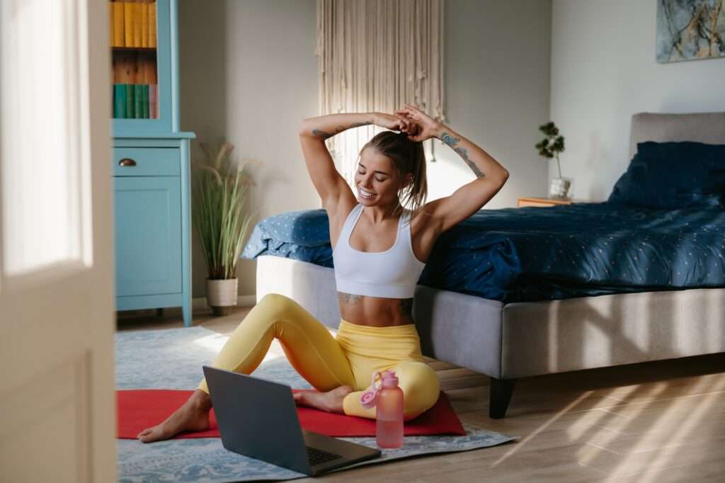 Happy sporty woman adjusting hair and looking at laptop while preparing to home workout